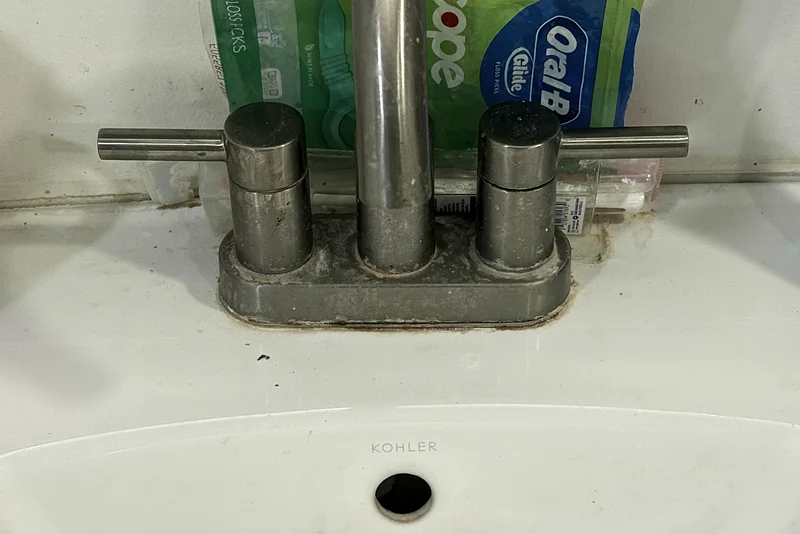 A brushed-nickel sink faucet before cleaning: hard-water scale, toothpaste splatter and mouthwash bottles crowding the counter