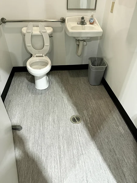 A commercial office restroom before cleaning: construction dust over the toilet, sink and vinyl floor, with a debris bin in the corner