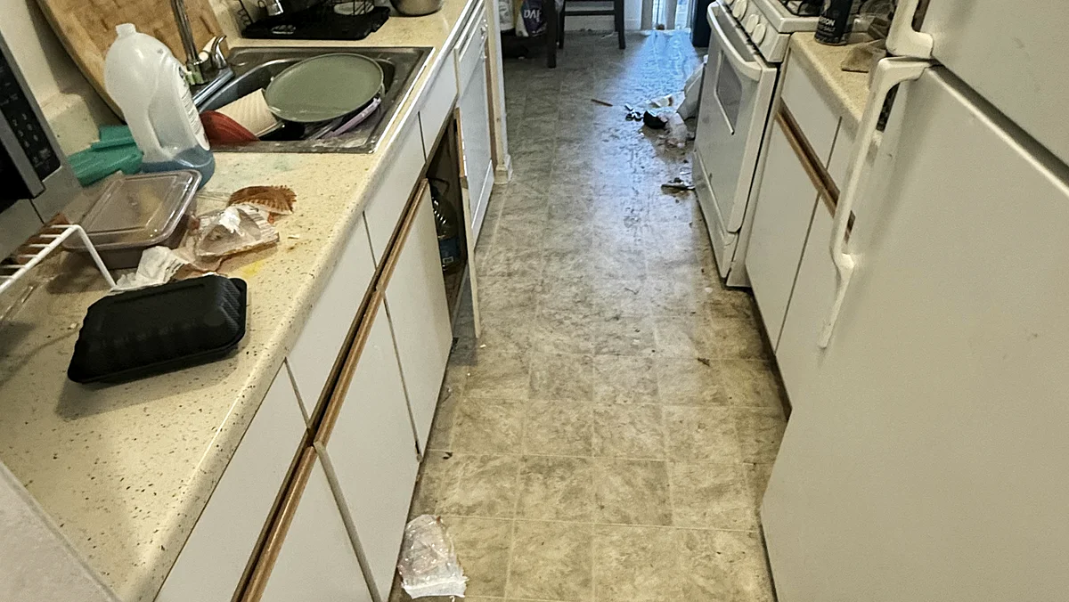 A galley apartment kitchen before cleaning: cluttered counters, food residue on the range and a dirty vinyl floor
